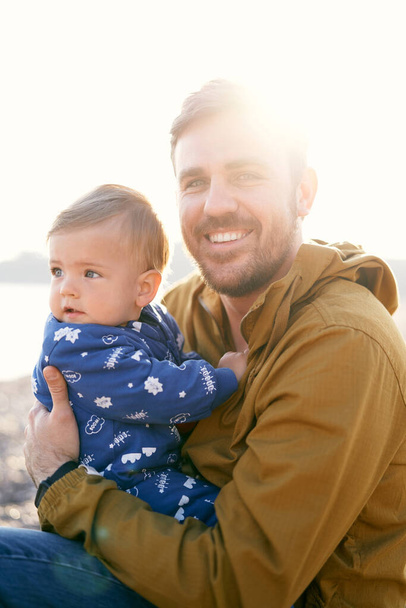 Улыбающийся папа в коричневой куртке сидит и держит маленького ребенка в синем комбинезоне на руках, повернутого боком при ярком солнечном свете. Крупный план - Фото, изображение