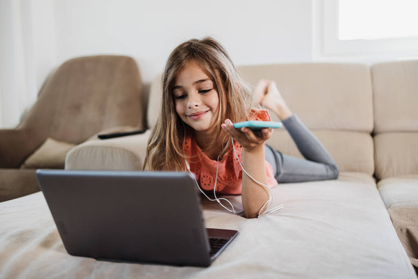 Beautiful girl lies on the living room sofa and uses a laptop for surfing, learning and communication. She is happy and smiled. - Фото, изображение