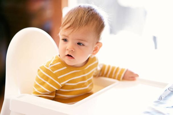 Słodkie dziecko siedzi na białym fotelu i odwraca wzrok. Zbliżenie - Zdjęcie, obraz
