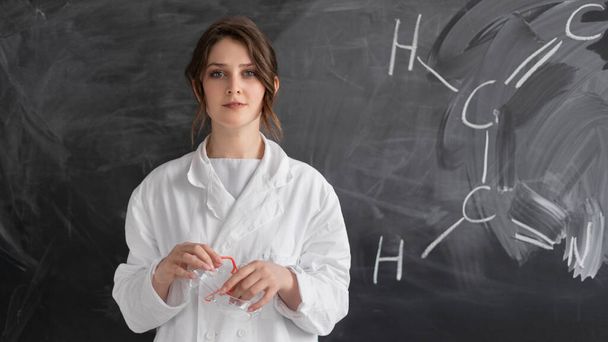Beyaz önlüklü beyaz bir kadın kimyasal formüllü bir tebeşir tahtasının arkasında duruyor. Kimya öğretmeni ya da özel öğretmen. Okula dönüş konsepti. Boşluğu kopyala. - Fotoğraf, Görsel