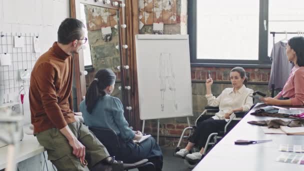 Mediana toma de diseñadora de moda femenina en silla de ruedas apuntando a la idea de la prenda dibujada en pizarra y conduciendo la reunión con su equipo en el estudio - Metraje, vídeo