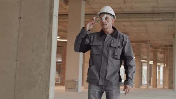Seguimiento del constructor masculino caucásico confiado de mediana edad que usa chaqueta de trabajador y sombrero blanco, caminando en el edificio en construcción, poniéndose gafas de seguridad - Imágenes, Vídeo