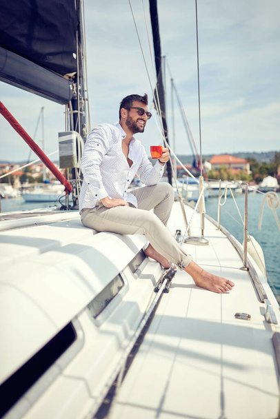 A young man sitting on a yacht and enjoying a coffe and a view on the dock on a beautiful sunny day. Summer, sea, vacation - Photo, Image
