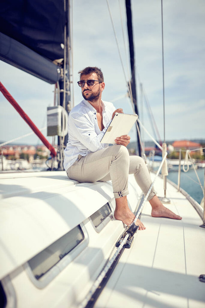 A young thoughtful businessman with a tablet is sitting on his yacht and watching a scenery while riding in the dock on a beautiful day on the seaside. Summer, sea, vacation - Fotoğraf, Görsel