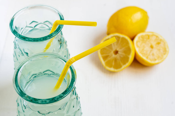 Vista superior de dos vasos con zumo de limón con paja amarilla y medio limones, enfoque selectivo, sobre mesa blanca, horizontal, con espacio para copiar - Foto, imagen