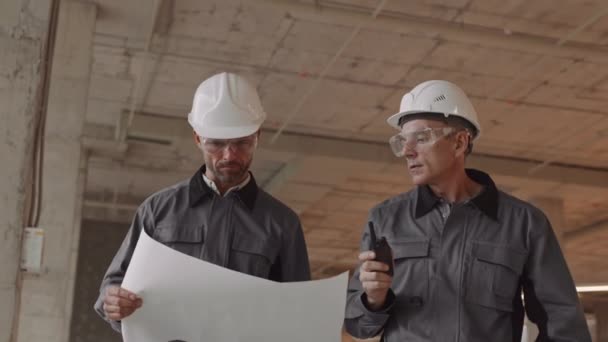Tracking shot of two Caucasian workers talking, wearing worker jackets, hard hats and safety goggles, walking around construction site, one man holding paper, his colleague - walkie-talkie - Footage, Video