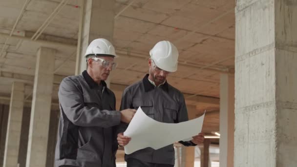 Kamerafahrt von zwei männlichen Kaukasiern in Arbeiterjacken, Harthüten und Schutzbrillen, die auf der Baustelle herumlaufen, ein Mann hält Papier, sein Kollege - Walkie-Talkie - Filmmaterial, Video