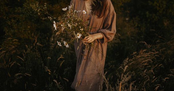 Mulher bonita em vestido de linho reunindo flores silvestres no prado de verão à noite. Imagem vintage elegante atmosférica. Elegante jovem mulher em vestido rústico colhendo flores no campo - Foto, Imagem