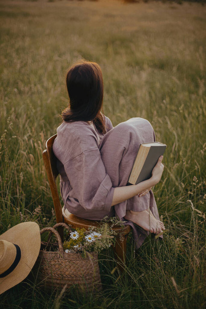 Belle femme en robe de lin assise sur une chaise rustique et profitant du coucher de soleil dans la prairie d'été. Jeune femme relaxante avec livre et panier de fleurs à la campagne. Moment de tranquillité atmosphérique - Photo, image