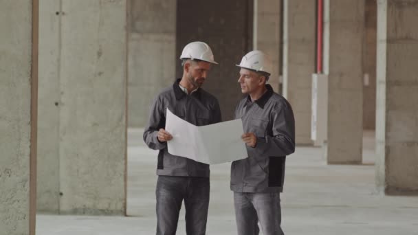 Středně dlouhý bělošský dělník a předák mluví, nosí pracovní vesty, tvrdé klobouky a bezpečnostní brýle, stojí na staveništi, drží papír s plánem projektu - Záběry, video
