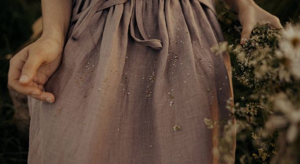 Belle robe en lin avec des pétales de fleurs sauvages sur la femme au milieu de l'herbe dans la prairie d'été, tissu près. Rassembler marguerite et persil de vache à la campagne. Atmosphérique image de rêve élégant - Photo, image