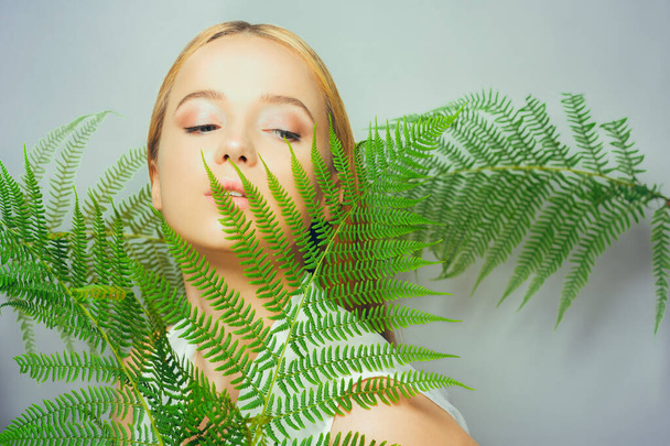 Beautiful young blond woman with perfect skin and natural make up posing front of plant tropical green leaves background. Teen model hair care of her face Fashion. - Fotoğraf, Görsel