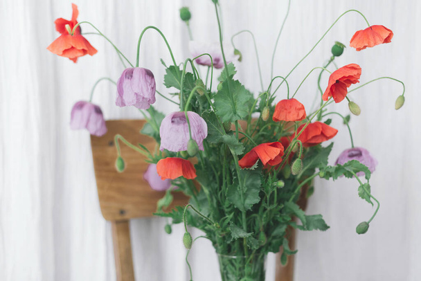 Beautiful red and purple poppies bouquet on wooden chair on background of rustic textile in room. Gathering countryside wildflowers. Common poppy and Opium poppy flowers in vase - Photo, Image