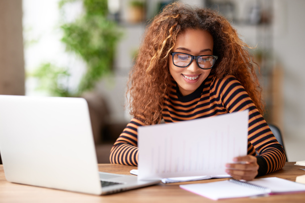 自宅のオフィスでドキュメントを操作しながら、ノートパソコンでリモートで作業しながら笑顔の明るい若いアフリカ系アメリカ人女性の肖像画。幸せな混合レース女性学生は自宅からオンラインで勉強 - 写真・画像
