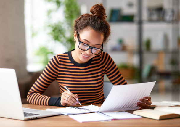 Όμορφη νεαρή Αφρο-Αμερικανίδα επιχειρηματίας που αναλύει έγγραφα ενώ κάθεται στο χώρο εργασίας της και εργάζεται εξ αποστάσεως σε φορητό υπολογιστή. Καλή μικτή φυλή φοιτήτρια που σπουδάζει online κατά τη διάρκεια της επιδημίας covid 19 - Φωτογραφία, εικόνα