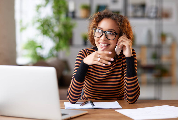 Wesoła młoda, afrykańska kobieta rozmawiająca z klientką przez telefon komórkowy, uśmiechająca się i robiąca notatki podczas zdalnej pracy na laptopie z domu, szczęśliwa pracownica biurowa dzwoniąca w interesach - Zdjęcie, obraz
