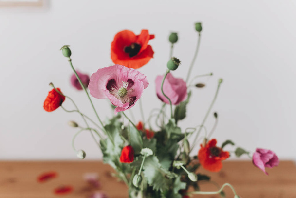 Beautiful poppies bouquet on wooden table in room, petals close up. Gathering countryside wildflowers, rural still life. Red common poppy and purple opium poppy flowers - Photo, Image