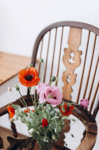 Beautiful poppies bouquet on rustic wooden chair in room. Gathering countryside wildflowers, rural still life. Red common poppy and purple opium poppy flowers in vase - Photo, Image