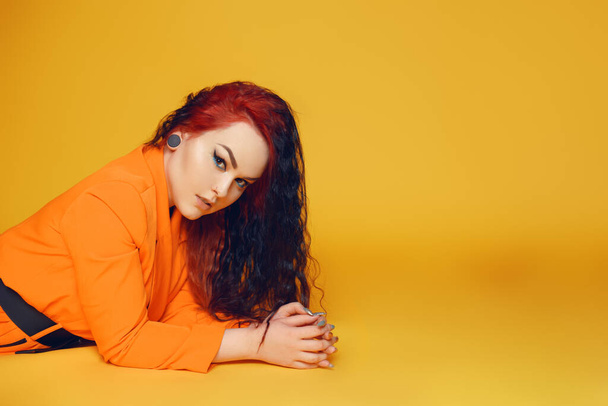 Egy hosszú, göndör hajú nő fekszik a padlón sárga háttér mellett. Egy lány narancssárga ruhában, vörös hajjal és nyilakkal a szemében. Divatos. Füldugó, fülbevaló. - Fotó, kép