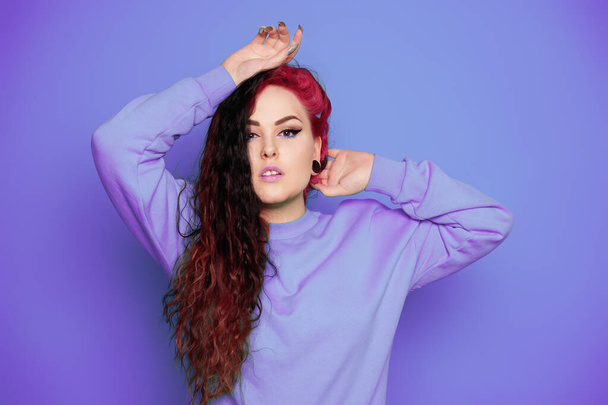 Woman in a blue sweater on a blue background posing. A beautiful brunette with long hair and and tunnels in her ears. Eyeliner and trendy makeup - Photo, Image