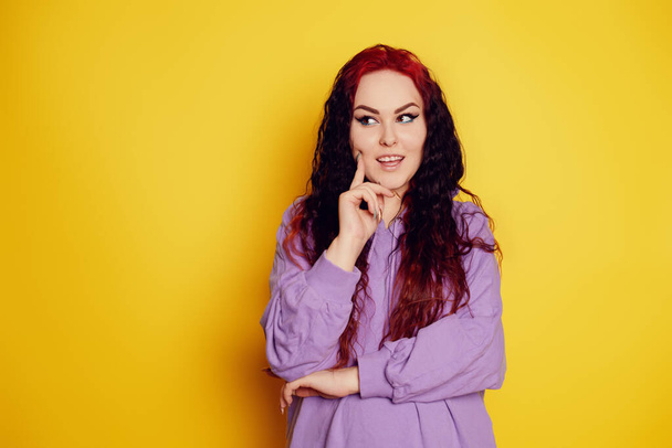 Woman in a purple sweater on a yellow background posing. A beautiful brunette with long curly hair and red roots. Model looks to the side and thinks, space for text, empty space - Photo, Image