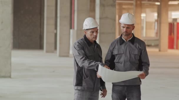 Lungo medio caposquadra caucasico che spiega il piano di progetto su carta ai dipendenti, uomini che indossano giacche da lavoro, cappelli duri e occhiali di sicurezza, in piedi in cantiere - Filmati, video