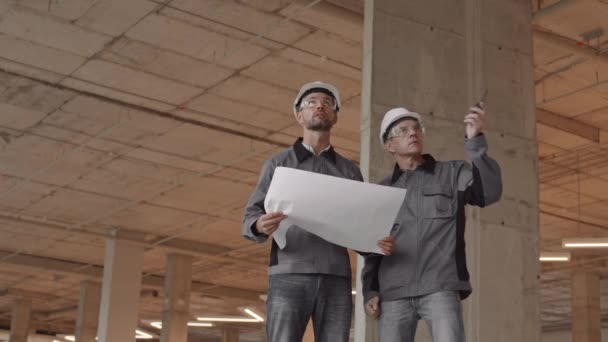 Medium long of two Caucasian workers talking, wearing worker jackets, hard hats and safety goggles, standing at construction site, one man holding paper, his colleague - walkie-talkie - Footage, Video