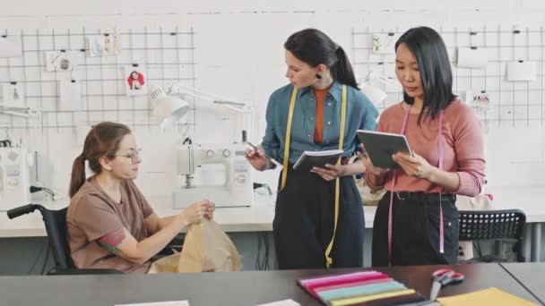 PAN-снимок женщин-дизайнеров с планшетом и ноутбуком, разговаривающих с швеей в инвалидной коляске, показывающий им одежду - Кадры, видео