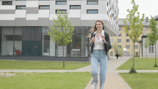 Young girl student, teenager, in a leather jacket with a backpack, stylishly dressed, Runs near a modern office or college building. Very Slow Movement, smile at the camera. Happy teenage girl enjoy - Foto, afbeelding