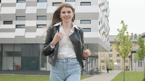 Young girl student, teenager, in a leather jacket with a backpack, stylishly dressed, Runs near a modern office or college building. Very Slow Movement, smile at the camera. Happy teenage girl enjoy - Photo, Image