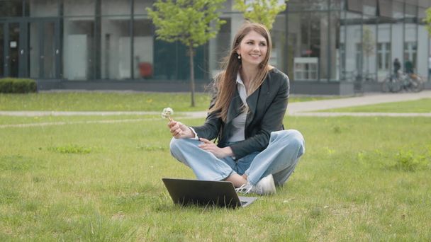 Молодая стильно одетая девушка сидит в парке на траве возле компьютера, держа одуванчик и дуя цветами. Медленное движение. Занятая девушка, работающая за ноутбуком, сидит на траве в городском парке - Фото, изображение