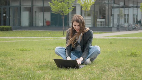 Joven chica elegantemente vestida sentada en el parque en la hierba cerca de la computadora, sosteniendo un diente de león y soplando flores. En cámara lenta. Chica ocupada trabajando en el portátil como sentado en la hierba en el parque de la ciudad - Foto, Imagen