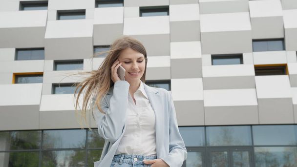 Кавказская уверенная молодая деловая женщина в белой рубашке разговаривает по телефону возле современного офисного здания. Medium Long Low Angle 4K Slow Motion Corporate Shot with Moving Around. Профессиональные люди - Фото, изображение