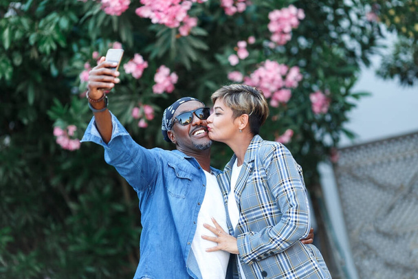 幸せな瞬間。ロマンチックなデート旅行をしながら、スマートフォンでセルフィーを取って陽気な黒のカップル,旅行中の中年アフリカ系アメリカ人配偶者.混合レースカップル  - 写真・画像