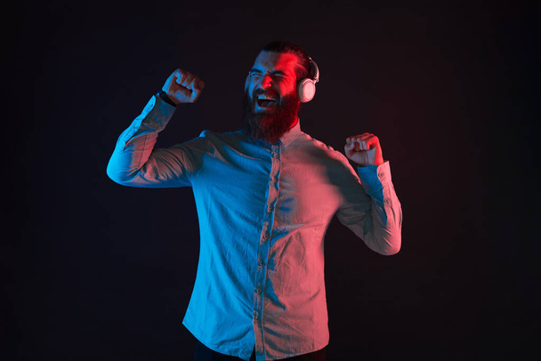 Foto de un joven barbudo hipster en música casual escuchando con las manos arriba sobre un fondo oscuro. - Foto, imagen