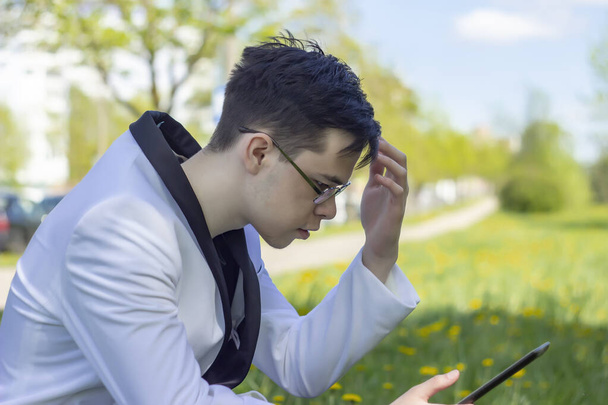 Pouliční portrét mladého muže ve věku 18-21 let s brýlemi, čtení informací z tabletu a posezení na trávě ve městě.Koncepce: začátek tréninku, kontrola kryptoměnového směnného kurzu. - Fotografie, Obrázek