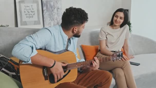 Média foto de jovem latino tocando músicas de guitarra para sua bela amada mulher ouvindo-o e batendo palmas sentados juntos no sofá no apartamento de design moderno - Filmagem, Vídeo