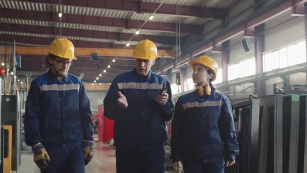 Medium slow mo van mannelijke volwassene ploegbaas met walkie-talkie in handen lopen door plant faciliteit praten met mannelijke en vrouwelijke fabrieksarbeiders in harde hoeden en overall - Video