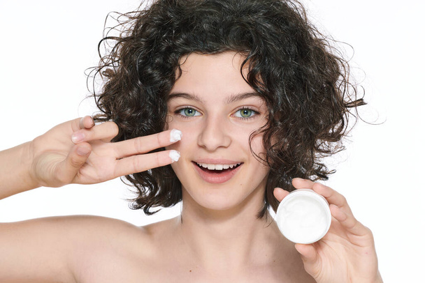 Teenager skincare. Beautiful teenage girl with gorgeous curly hair holding moisturiser face cream, looking at camera smiling. Studio shot on white background. Beauty, skincare and puberty concept. - Foto, Bild