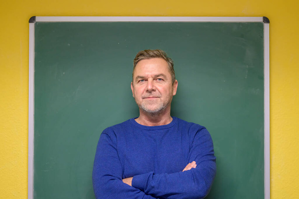 Serieuze mannelijke leraar staat met gevouwen armen voor een schoon groen schoolbord op een muur in een klaslokaal - Foto, afbeelding