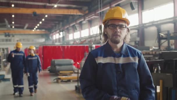 Střední PAN portrét s pomalým pózováním továrního dělníka pro kameru se zkříženýma rukama, zatímco jeho mužští a ženští kolegové procházejí továrnou v pozadí - Záběry, video