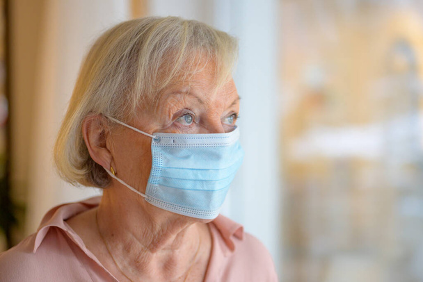 Donna anziana che indossa una maschera chirurgica monouso durante l'isolamento per la pandemia di Coronavirus in piedi guardando pensieroso fuori da una finestra - Foto, immagini