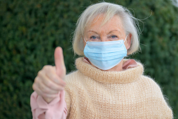 Положительная пожилая женщина подает большой палец вверх, чтобы показать, что все хорошо, как она стоит на улице в зимнем саду в защитной маске от коронавируса во время пандемии - Фото, изображение