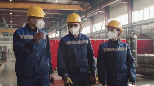 Média lentidão do supervisor de turnos com trabalhadores masculinos e femininos em máscaras respiratórias, chapéus duros e uniformes andando pelas instalações da planta e conversando - Filmagem, Vídeo