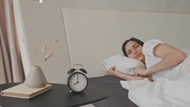 Средний снимок сонной молодой женщины латинской национальности, лежащей в постели и потревоженной звонящим будильником на тумбочке, выключающим ее и продолжающим спать - Кадры, видео