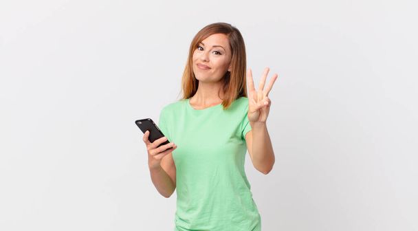 pretty woman smiling and looking friendly, showing number three and using a smartphone - Фото, зображення