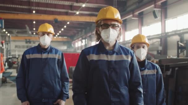 Keskikokoinen muotokuva tehtaan johtajasta ja kahdesta työpaikalla seisovasta mies- ja naispuolisesta työntekijästä hengityssuojaimissa, kypärissä ja haalareissa, jotka poseeraavat kameralle - Materiaali, video