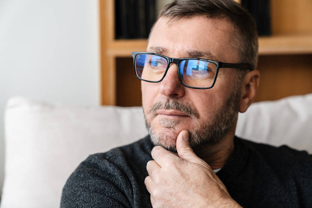 Serious adult unshaven man in eyeglasses looking aside while sitting indoors - Foto, Bild