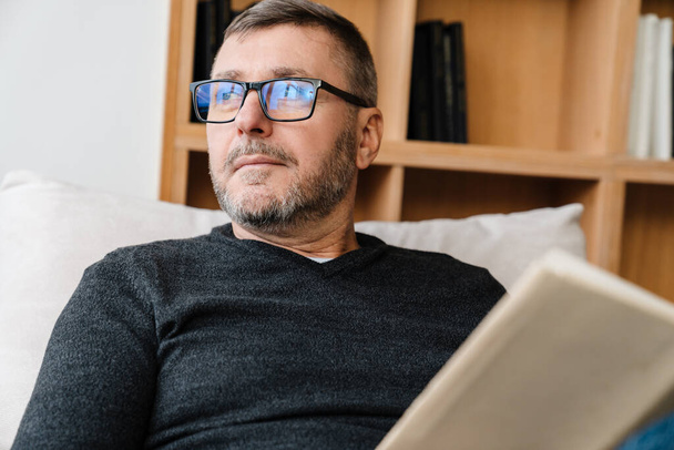 Serious adult unshaven man in eyeglasses reading book while resting at home - Foto, immagini