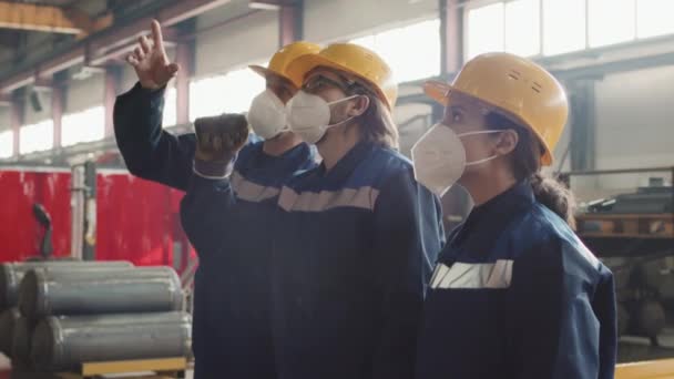 Média lentidão do supervisor masculino explicando novos trabalhadores masculinos e femininos como operar máquinas na fábrica, todos usando máscaras respiratórias, chapéus duros e uniformes - Filmagem, Vídeo